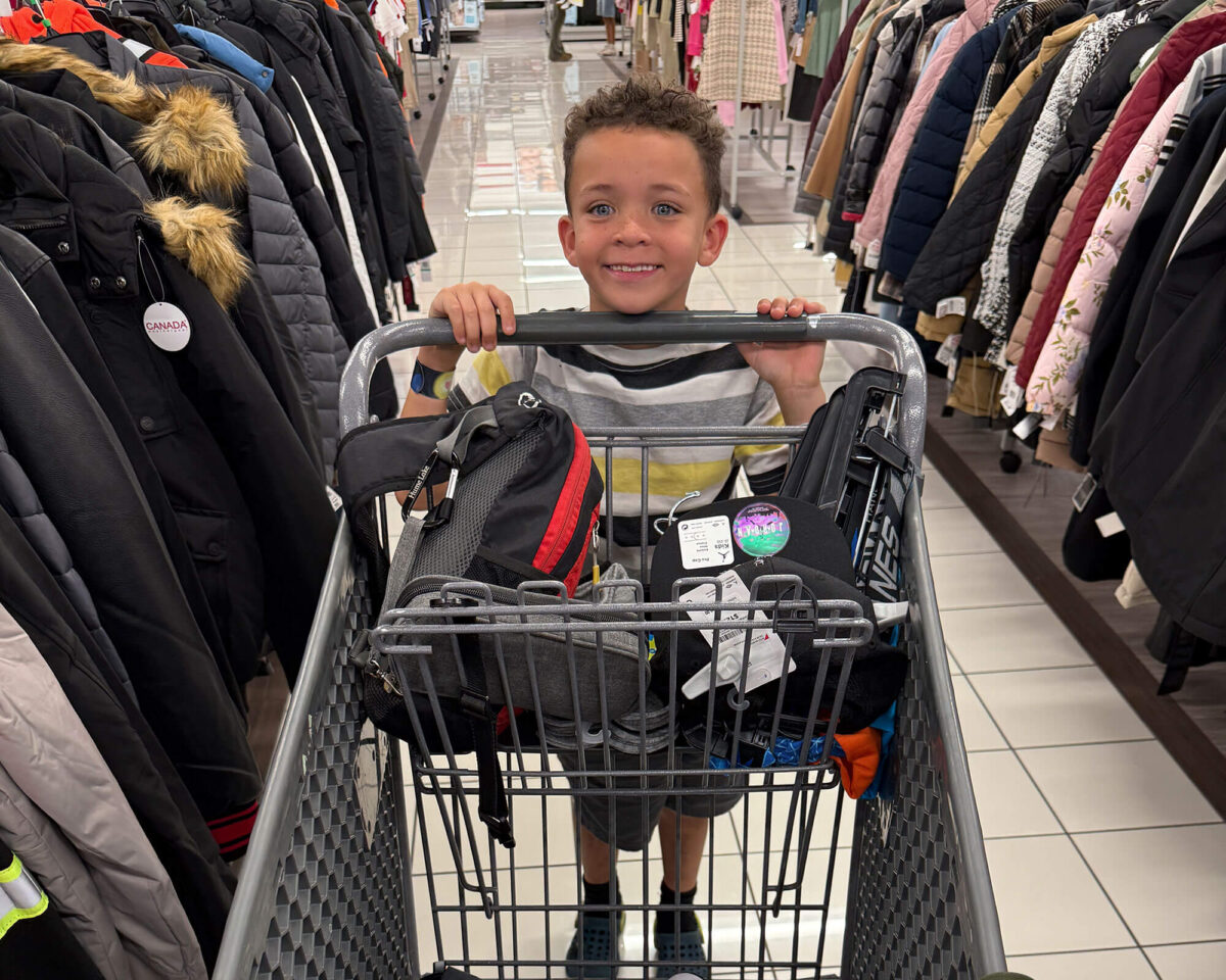 Smiling young boy seated in a shopping cart filled with new backpacks at a retail clothing store
