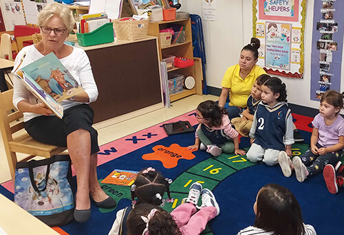 Volunteer reading a picture book aloud to preschool children seated on a colorful classroom rug