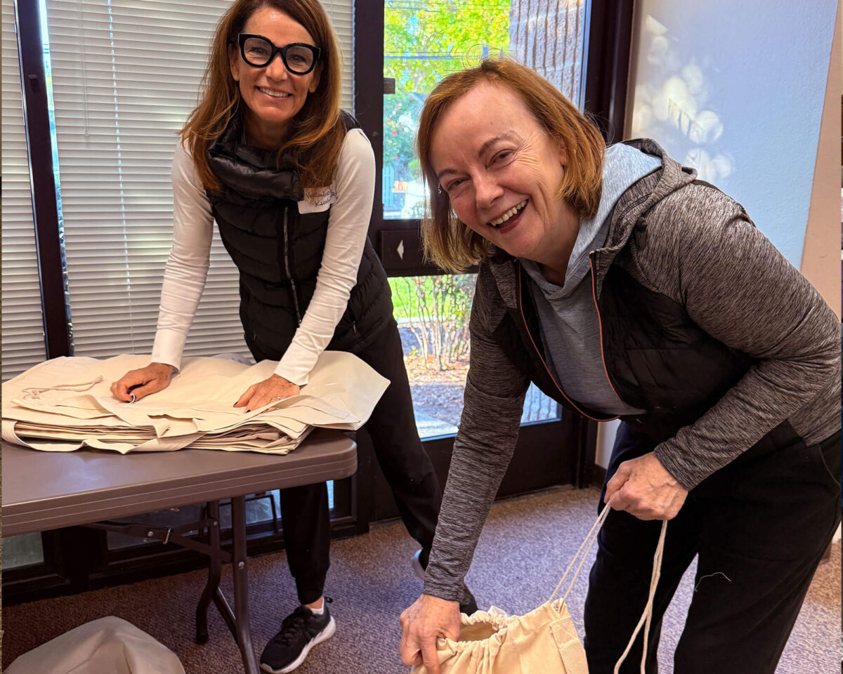 Two volunteers laughing while filling drawstring cloth care packs at a table