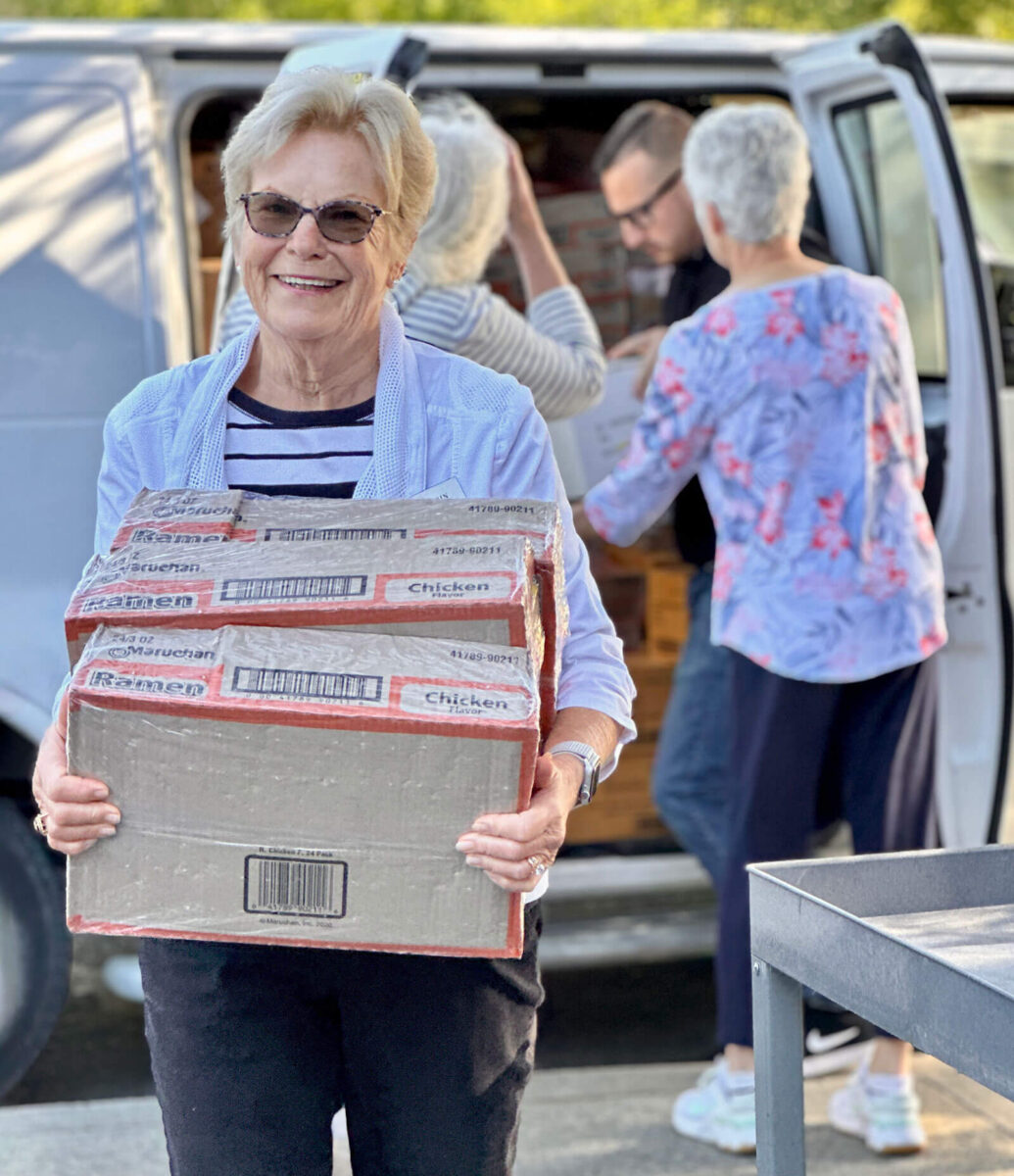 Volunteer smiling while carrying boxes of ramen as fellow volunteers load a delivery van behind her