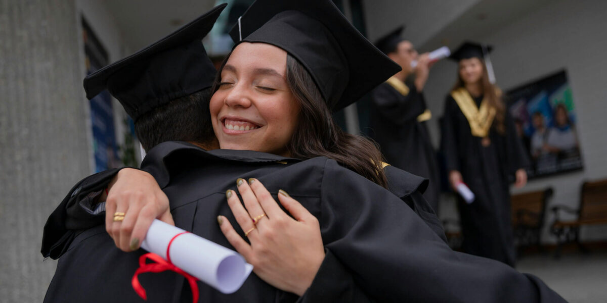 Two graduates in caps and gowns embracing while holding their diplomas at a graduation ceremony