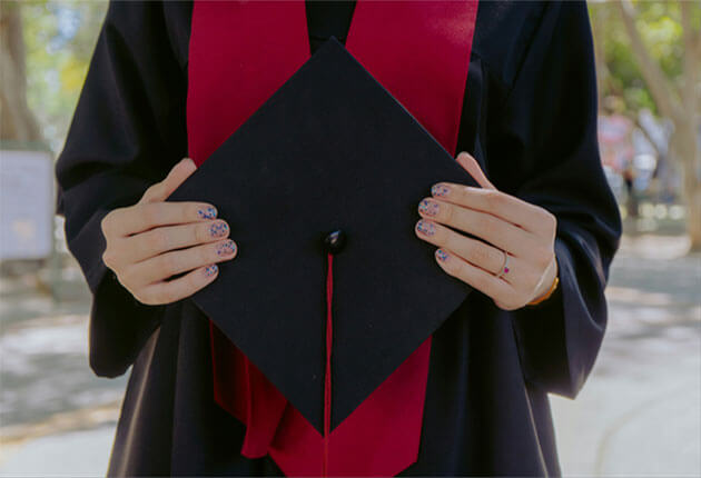 Graduate holding their mortarboard cap in front of a black gown with red sash