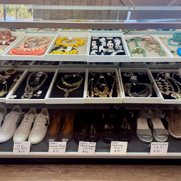 Display case at the Giving Partners Thrift Shop featuring trays of jewelry above a row of priced gently-used shoes