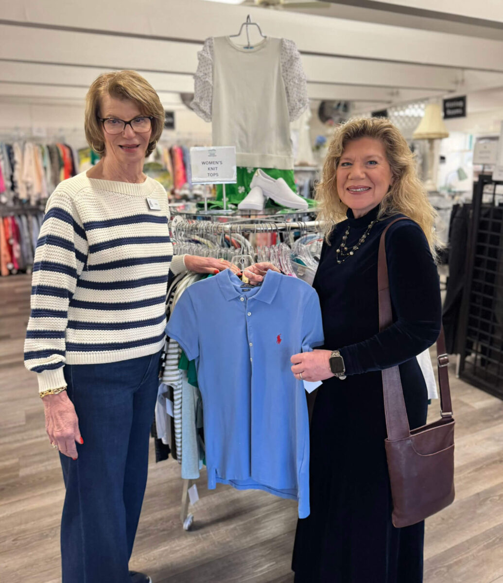 Two volunteers showing a gently-used Ralph Lauren polo shirt at the Giving Partners Thrift Shop's women's knit tops display