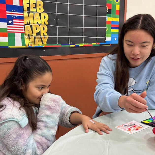 Volunteen face-painting a young child's hand at a community event craft table
