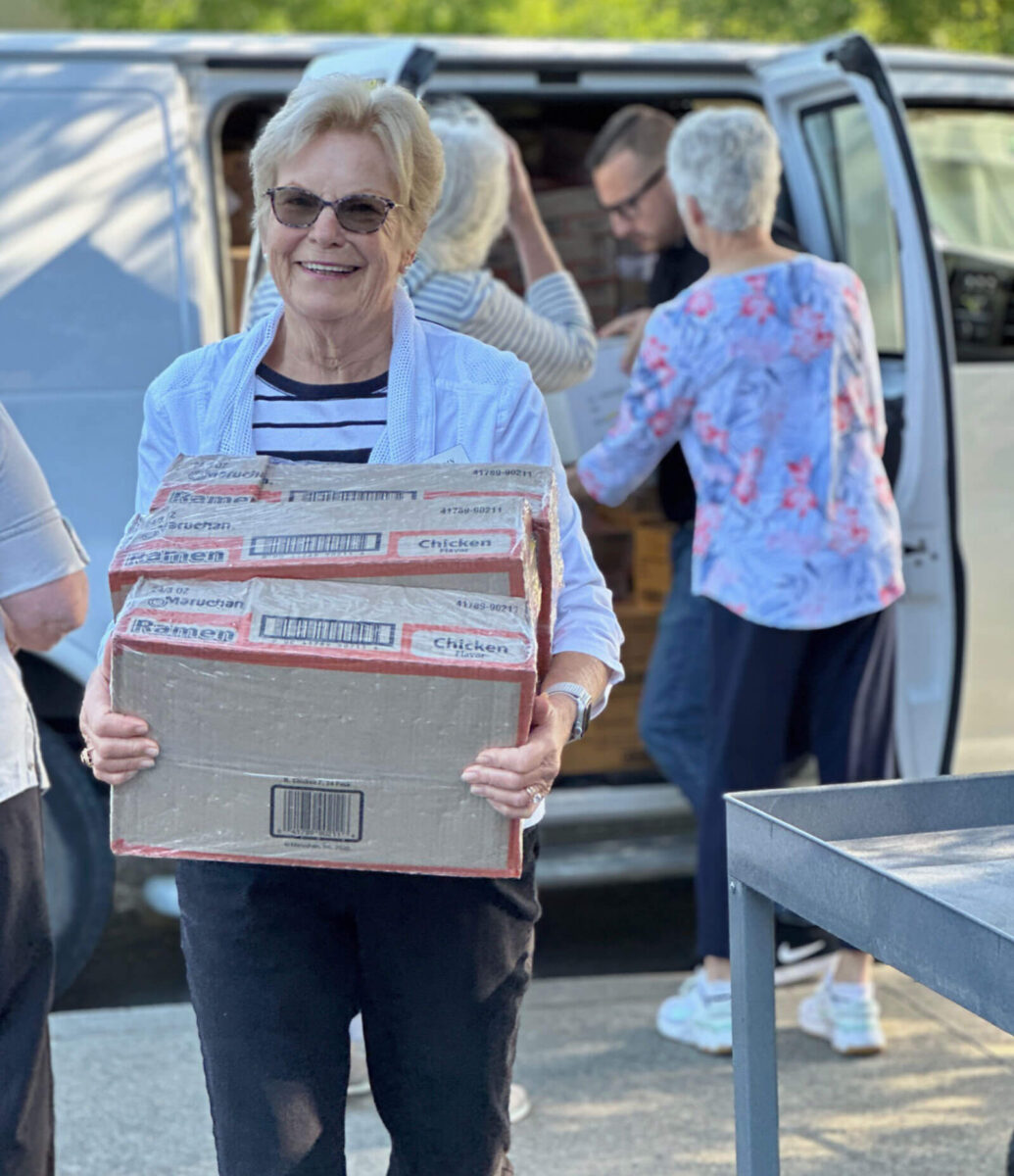 Volunteer smiling while carrying boxes of ramen as fellow volunteers load a delivery van behind her